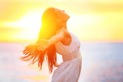 Enjoyment - free happy woman enjoying sunset. Beautiful woman in a white dress embracing the golden sunshine glow of sunset with her arms outspread and face raised to the sky enjoying peace and serenity of nature